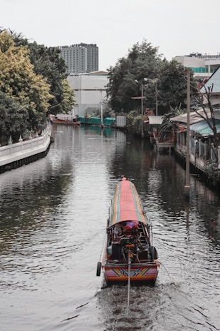 A long-tail boat travels down a narrow canal flanked by trees and buildings. The water reflects the colors of the environment, creating a serene setting. The background features urban structures, including a tall building.
