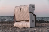 A woven beach chair stands on a sandy beach with a backdrop of calm ocean waters and a serene sky. The chair is predominantly white with red accents, and its design features rounded, protective sides.