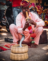 A man wearing a colorful turban and a light pink outfit is playing a traditional musical instrument, a pungi, seated on the ground. In front of him, a snake is emerging from a wicker basket. The background includes vibrant textiles and a parked scooter.