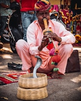 A man wearing a colorful turban and a light pink outfit is playing a traditional musical instrument, a pungi, seated on the ground. In front of him, a snake is emerging from a wicker basket. The background includes vibrant textiles and a parked scooter.
