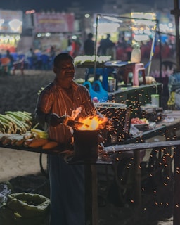 A street vendor is roasting corn over an open flame, with sparks flying around. The background is bustling with various food stalls and colorful lights in a market setting during the night.