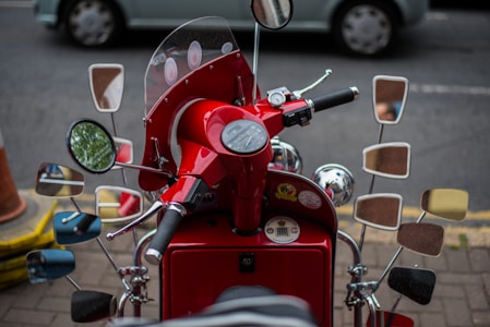 A red vintage scooter adorned with numerous mirrors on both sides of the handlebars, providing a unique and retro appearance. The dashboard displays gauges including a speedometer, and the background shows a parked car on a street.