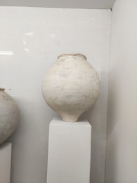 A large, rounded clay pot with a smooth surface is displayed on a white pedestal in a well-lit room. The pot has a light color with subtle markings and imperfections, suggesting it is hand-made or aged.