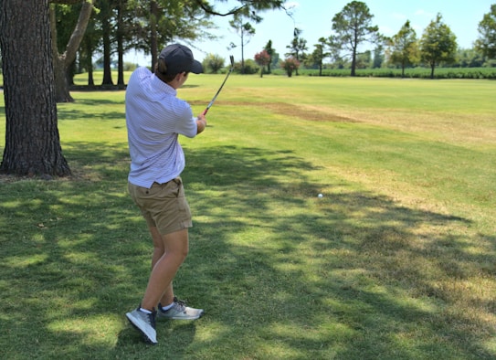 A person wearing a cap, a striped short-sleeve shirt, and khaki shorts is swinging a golf club in a grassy area surrounded by trees. The landscape includes a well-maintained golf course with green grass and a few trees lining the horizon under a clear sky.