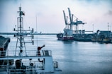 A waterfront industrial area with a large cargo ship docked near towering cranes, surrounded by a calm body of water. The foreground features the upper structure of a ship, including communication antennas and other equipment. The industrial infrastructure on the shore is dense with buildings and machinery.
