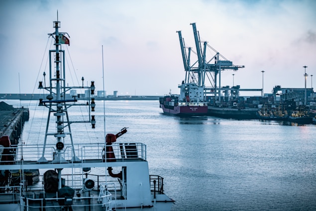 A waterfront industrial area with a large cargo ship docked near towering cranes, surrounded by a calm body of water. The foreground features the upper structure of a ship, including communication antennas and other equipment. The industrial infrastructure on the shore is dense with buildings and machinery.