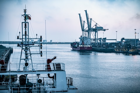 A waterfront industrial area with a large cargo ship docked near towering cranes, surrounded by a calm body of water. The foreground features the upper structure of a ship, including communication antennas and other equipment. The industrial infrastructure on the shore is dense with buildings and machinery.