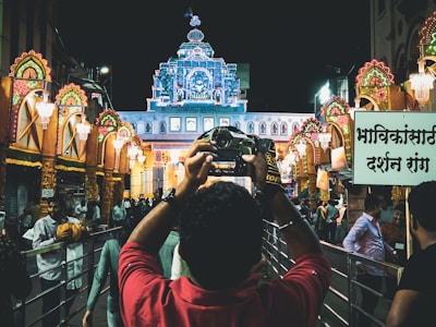 A vibrant night scene captures a man taking a photograph of an ornately decorated building illuminated with colorful lights. The street is bustling with people, and the architecture is adorned with intricate details and bright lights. Traditional elements and festive decorations are prominent, creating a lively atmosphere.