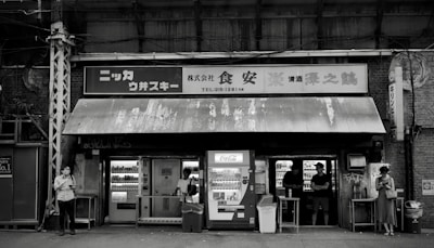 A black and white image of a small, weathered convenience store with vintage signage and vending machines. There are multiple people standing outside, some interacting with the machines and others using their phones. The store has an assortment of shelves visible through the windows.