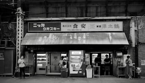 A black and white image of a small, weathered convenience store with vintage signage and vending machines. There are multiple people standing outside, some interacting with the machines and others using their phones. The store has an assortment of shelves visible through the windows.