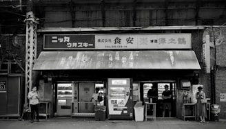 A black and white image of a small, weathered convenience store with vintage signage and vending machines. There are multiple people standing outside, some interacting with the machines and others using their phones. The store has an assortment of shelves visible through the windows.