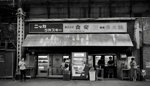 A black and white image of a small, weathered convenience store with vintage signage and vending machines. There are multiple people standing outside, some interacting with the machines and others using their phones. The store has an assortment of shelves visible through the windows.