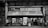 A black and white image of a small, weathered convenience store with vintage signage and vending machines. There are multiple people standing outside, some interacting with the machines and others using their phones. The store has an assortment of shelves visible through the windows.