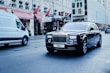 A luxurious black car with a custom license plate drives down a city street. Nearby, a white commercial van is also on the move. The background features an elegant building with red awnings and visible signage, indicating a busy urban environment.