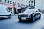 A luxurious black car with a custom license plate drives down a city street. Nearby, a white commercial van is also on the move. The background features an elegant building with red awnings and visible signage, indicating a busy urban environment.