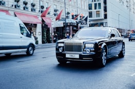 A luxurious black car with a custom license plate drives down a city street. Nearby, a white commercial van is also on the move. The background features an elegant building with red awnings and visible signage, indicating a busy urban environment.