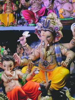 A variety of colorful Ganesha idols are displayed together. Each statue features detailed ornaments, with vibrant colors like yellow, red, and blue. The Ganesha figures are depicted in various poses and adorned with jewelry.