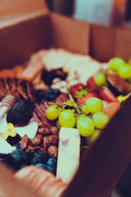 A close-up of a charcuterie box containing a variety of foods. There are visible grapes on a vine, blueberries, sliced meats, cheeses, nuts, and other fruits and snacks. The box is made of brown cardboard, and the items are neatly arranged inside.