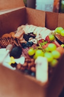 A cardboard box filled with a variety of fresh fruits, nuts, and snacks. Green grapes, strawberries, blueberries, and blackberries are visible alongside crackers, mixed nuts, and slices of bread. The assortment appears appetizing and colorful.