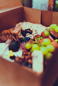 A cardboard box filled with a variety of fresh fruits, nuts, and snacks. Green grapes, strawberries, blueberries, and blackberries are visible alongside crackers, mixed nuts, and slices of bread. The assortment appears appetizing and colorful.