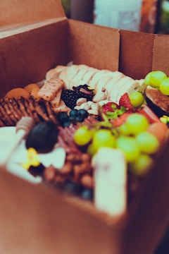 A cardboard box filled with a variety of fresh fruits, nuts, and snacks. Green grapes, strawberries, blueberries, and blackberries are visible alongside crackers, mixed nuts, and slices of bread. The assortment appears appetizing and colorful.