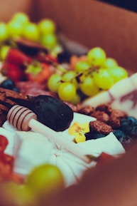 A vibrant assortment of fresh fruits and nuts arranged in a decorative manner, featuring grapes, berries, and assorted nuts accompanied by a floral garnish and a honey dipper.
