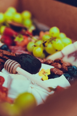 A vibrant assortment of fresh fruits and nuts arranged in a decorative manner, featuring grapes, berries, and assorted nuts accompanied by a floral garnish and a honey dipper.