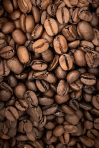A close-up view of roasted coffee beans piled together, showcasing their rich brown color and various sizes. The beans have visible textures and natural grooves, indicating their quality and roast level.