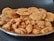 A metal plate filled with two types of golden-brown snacks is placed on a dark surface. At the back of the plate, there are round, slightly puffy fried disks, possibly a type of savory biscuit or cracker. In the front, curly, textured snacks similar to fritters or deep-fried bites are visible. Both snacks appear crispy and well-seasoned.