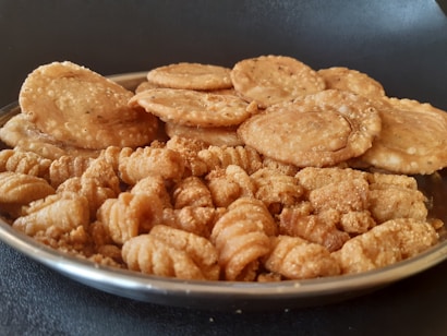 A metal plate filled with two types of golden-brown snacks is placed on a dark surface. At the back of the plate, there are round, slightly puffy fried disks, possibly a type of savory biscuit or cracker. In the front, curly, textured snacks similar to fritters or deep-fried bites are visible. Both snacks appear crispy and well-seasoned.