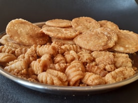 A metal plate filled with two types of golden-brown snacks is placed on a dark surface. At the back of the plate, there are round, slightly puffy fried disks, possibly a type of savory biscuit or cracker. In the front, curly, textured snacks similar to fritters or deep-fried bites are visible. Both snacks appear crispy and well-seasoned.