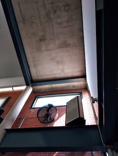 A modern architectural interior featuring a high ceiling with concrete panels and a wall made of red bricks. There is a ceiling fan mounted on the wall and a large rectangular speaker. A window is positioned above, allowing natural light to enter the space.