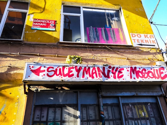 A building facade with a bright yellow wall and windows. There are signs in various languages, including one with red lettering pointing towards the Suleymaniye Mosque. The windows display colorful hanging chains, and the building appears to be situated in an urban setting.