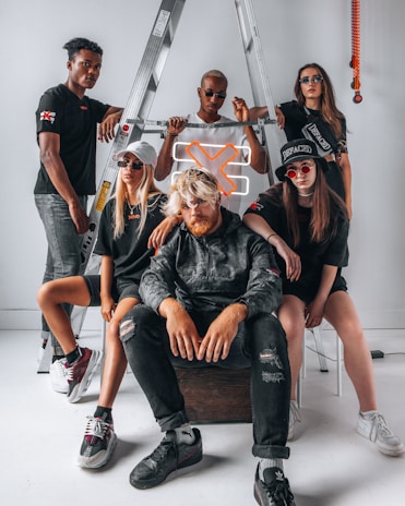 A group of five individuals pose confidently in front of a metal ladder. They are wearing stylish streetwear, including dark sunglasses and casual shoes. A neon orange 'X' sign is incorporated into the setting, adding a modern touch. The group displays various expressions and postures that suggest both unity and individuality.