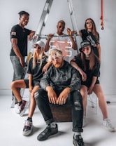 A group of five individuals pose confidently in front of a metal ladder. They are wearing stylish streetwear, including dark sunglasses and casual shoes. A neon orange 'X' sign is incorporated into the setting, adding a modern touch. The group displays various expressions and postures that suggest both unity and individuality.