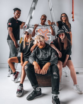A group of five individuals pose confidently in front of a metal ladder. They are wearing stylish streetwear, including dark sunglasses and casual shoes. A neon orange 'X' sign is incorporated into the setting, adding a modern touch. The group displays various expressions and postures that suggest both unity and individuality.