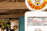 A vintage gasoline pump is prominently displayed, featuring a circular emblem with the word 'GASOLINE' in bold red letters against a yellow background. The pump's glass display shows 'REGULAR' in red letters, and a small metal indicator is visible beneath it. In the background, there is a structure with a truss roof and a sign for 'Goodyear Tires' in blue and yellow.