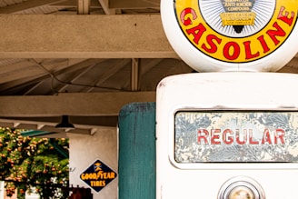 A vintage gasoline pump is prominently displayed, featuring a circular emblem with the word 'GASOLINE' in bold red letters against a yellow background. The pump's glass display shows 'REGULAR' in red letters, and a small metal indicator is visible beneath it. In the background, there is a structure with a truss roof and a sign for 'Goodyear Tires' in blue and yellow.