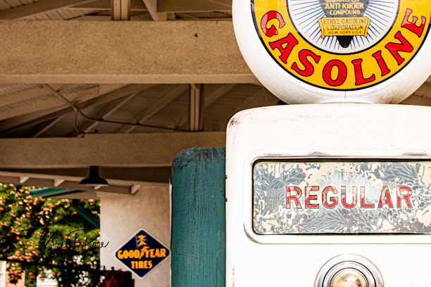 A vintage gasoline pump is prominently displayed, featuring a circular emblem with the word 'GASOLINE' in bold red letters against a yellow background. The pump's glass display shows 'REGULAR' in red letters, and a small metal indicator is visible beneath it. In the background, there is a structure with a truss roof and a sign for 'Goodyear Tires' in blue and yellow.