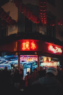 Neon lights illuminate an urban street scene featuring a busy storefront. Red and yellow neon signs, some with Chinese characters, create a vibrant atmosphere. Several people are gathered near the entrance, suggesting a lively and energetic environment.