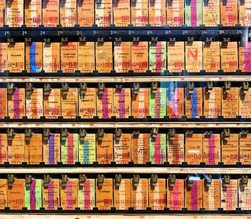 A collection of vintage train tickets displayed in rows, each ticket held in place by a metal clip. They feature various shades of orange, yellow, and blue with printed text denoting different destinations and travel information. Some tickets have bold, colorful highlights in pink and green, adding contrast to the overall muted tones.
