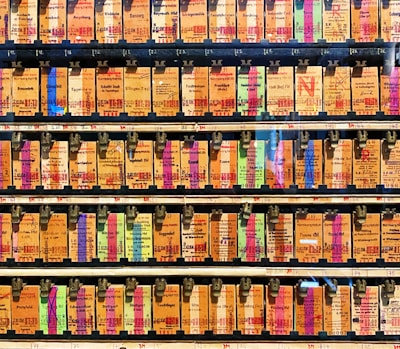 A collection of vintage train tickets displayed in rows, each ticket held in place by a metal clip. They feature various shades of orange, yellow, and blue with printed text denoting different destinations and travel information. Some tickets have bold, colorful highlights in pink and green, adding contrast to the overall muted tones.