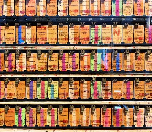 A collection of vintage train tickets displayed in rows, each ticket held in place by a metal clip. They feature various shades of orange, yellow, and blue with printed text denoting different destinations and travel information. Some tickets have bold, colorful highlights in pink and green, adding contrast to the overall muted tones.