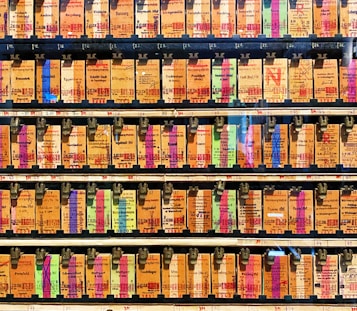 A collection of vintage train tickets displayed in rows, each ticket held in place by a metal clip. They feature various shades of orange, yellow, and blue with printed text denoting different destinations and travel information. Some tickets have bold, colorful highlights in pink and green, adding contrast to the overall muted tones.