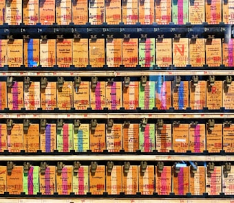 A collection of vintage train tickets displayed in rows, each ticket held in place by a metal clip. They feature various shades of orange, yellow, and blue with printed text denoting different destinations and travel information. Some tickets have bold, colorful highlights in pink and green, adding contrast to the overall muted tones.