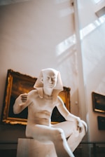 A marble sculpture of an ancient Egyptian figure is prominently displayed in a museum setting. The figure is seated and has a traditional Egyptian headdress. The background includes framed artwork on the walls, with soft natural light streaming in from above, creating a serene atmosphere.