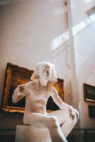 A marble sculpture of an ancient Egyptian figure is prominently displayed in a museum setting. The figure is seated and has a traditional Egyptian headdress. The background includes framed artwork on the walls, with soft natural light streaming in from above, creating a serene atmosphere.