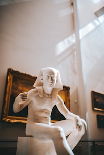 A marble sculpture of an ancient Egyptian figure is prominently displayed in a museum setting. The figure is seated and has a traditional Egyptian headdress. The background includes framed artwork on the walls, with soft natural light streaming in from above, creating a serene atmosphere.
