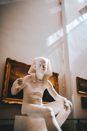 A marble sculpture of an ancient Egyptian figure is prominently displayed in a museum setting. The figure is seated and has a traditional Egyptian headdress. The background includes framed artwork on the walls, with soft natural light streaming in from above, creating a serene atmosphere.