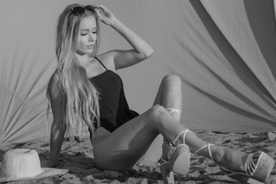 A woman with long hair sits on a sandy beach, wearing a dark one-piece swimsuit and strappy platform sandals. She appears relaxed, with one hand adjusting her sunglasses. Beside her is a straw hat resting on the sand, and in the background, a fabric structure or tent provides shade.